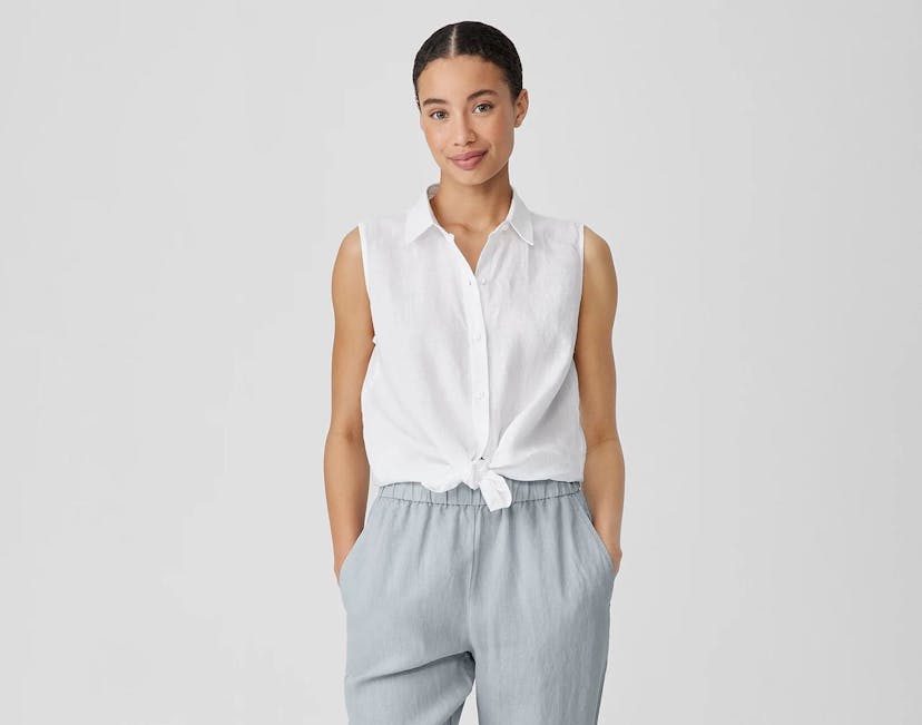 A woman with her hair pulled back wears a white, sleeveless linen blouse with a knot tied in front of the stomach and grey linen pants. She has her hands tucked in the pockets.