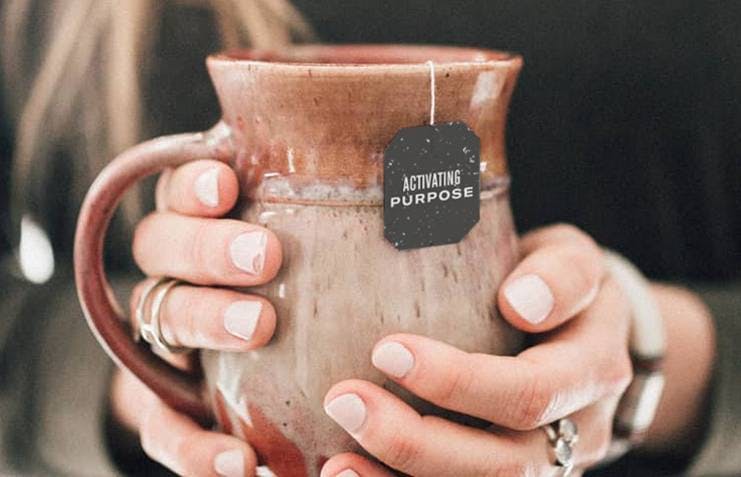 A pair of woman's hands holding a brown ceramic coffee mug, showing a tea bag label reading "Activate Purpose"