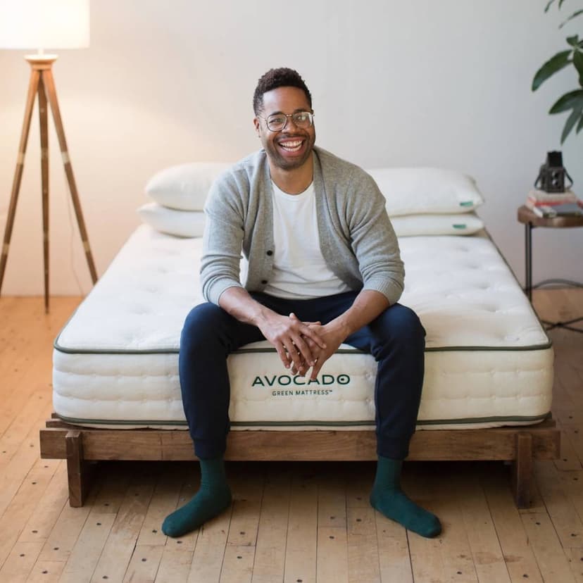 A smiling man sits on a mattress with his feet on the floor and his hands folded between his knees. The room has light wood floors and a tall lamp and a plant for decoration.