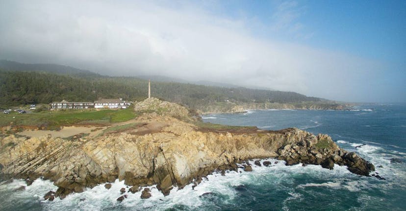 An aerial photo shows a view of the rocky coastline of California’s Sonoma County. A wide, two-story structure is visible, a white building with brown roof and trim. The building is the Timber Cove Resort.
