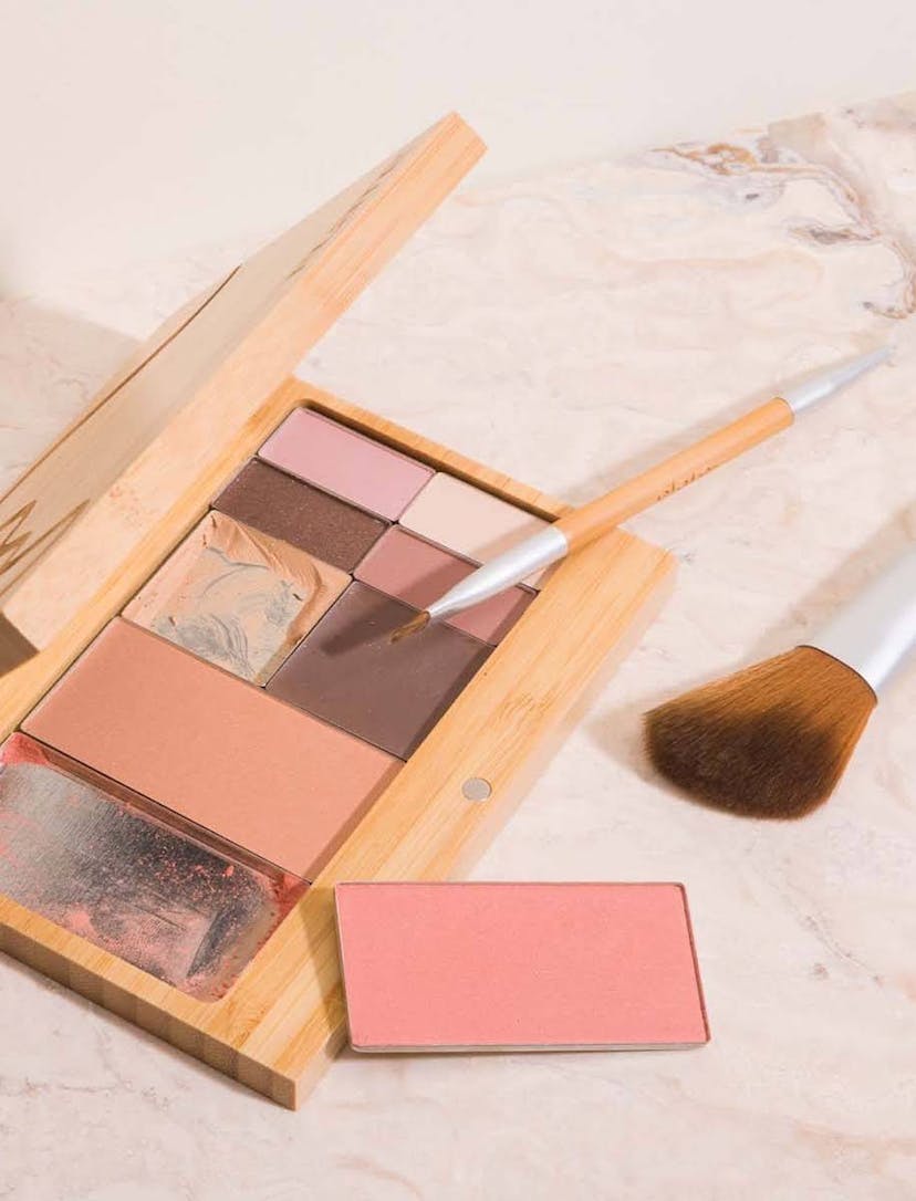 On a marble surface is an open, used bamboo Capsule Palette from Elate Beauty. It contains different shades of powder blush, eyeshadow, and eyeliner. A large face brush and small eyeshadow brush lie near the palette, which can be refilled with products of the consumer's choosing. Photo credit - Elate Beauty