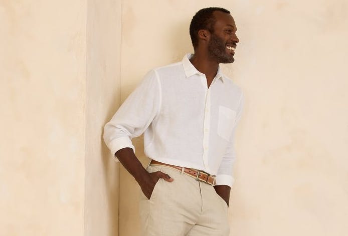 A relaxed-looking man smiles, wearing a pair of khaki trousers and a cream-colored linen button-up shirt from Tommy Bahama.