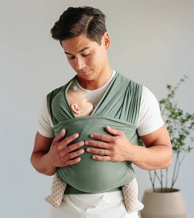 A person carries a baby in a green, wrap-style carrier.