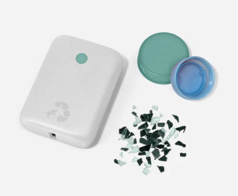 A small white portable charger with the recycling logo and a small elephant on it is shown next to two plastic bottle caps and shards of broken plastic.