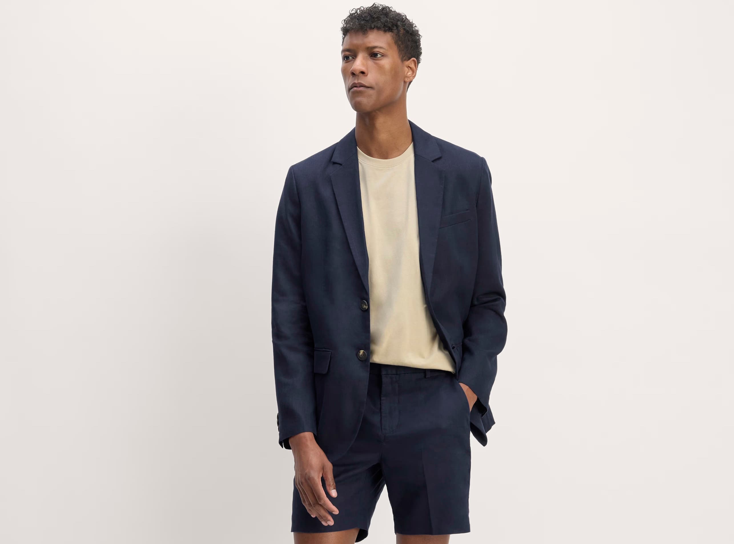 A man wears a suit of navy linen shorts and a navy linen blazer and a beige tee shirt.