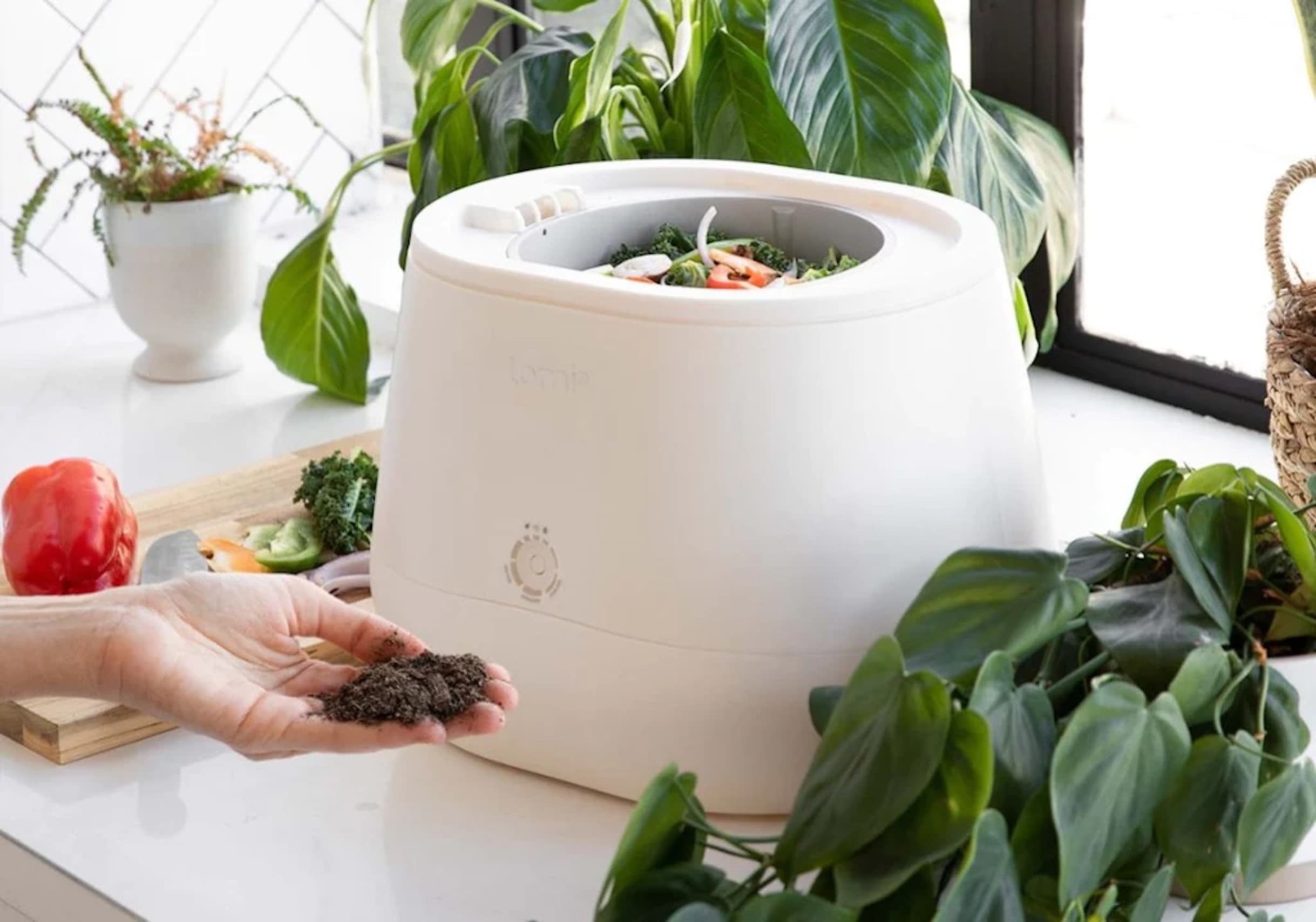 A small white machine with an open top has vegetable scraps in it and is shown on a kitchen countertop, with a cutting board and vegetables nearby. In front of the machine, a person's hand holds dirt.