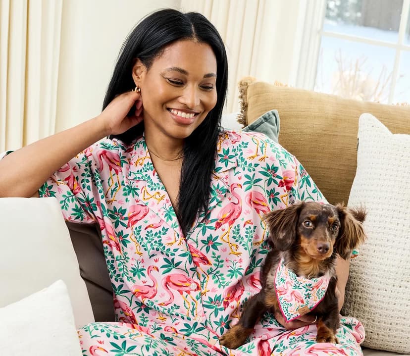 A smiling woman sits on a white sofa holding a small brown dog. The woman wears pink pajamas with a flamingo on them, and the dog wears a matching bandana.