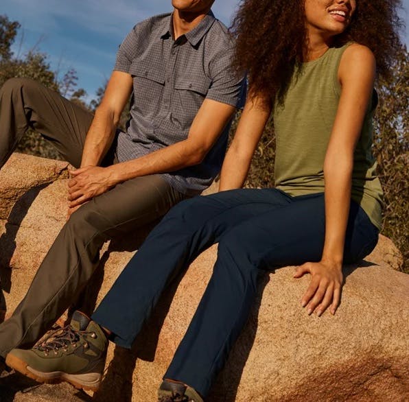 Two people sit on a rock in scrubby wilderness wearing outdoor pants and shirts appropriate for warm weather.