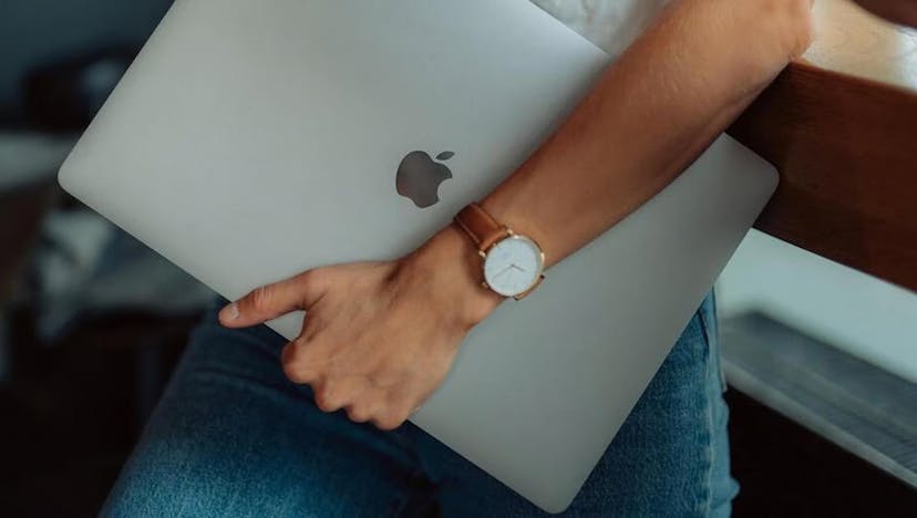 A person is shown from knee to elbow, walking while carrying a Macbook laptop tucked under the arm. The person, in bluejeans and a tee-shirt, wears a simple watch on a brown leather strap. Photo courtesy Back Market.