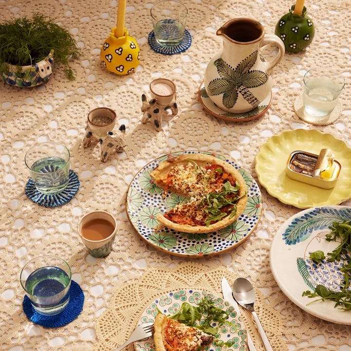 A table with a traditional cream-colored lace tablecloth is set with attractive hand-painted dishes, vases, jugs, and candleholders.