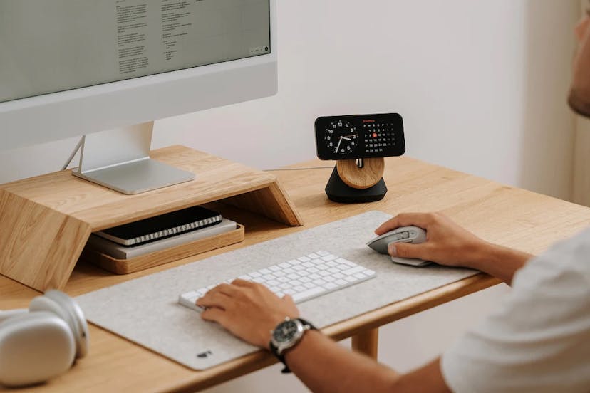 A man sits at a light blond wood desk, with his hands on a white Apple keyboard over a grey felt desk mat. Matching wooden desk accessories elevate his Apple desktop computer and his iPhone. Photo courtesy Oakywood.
