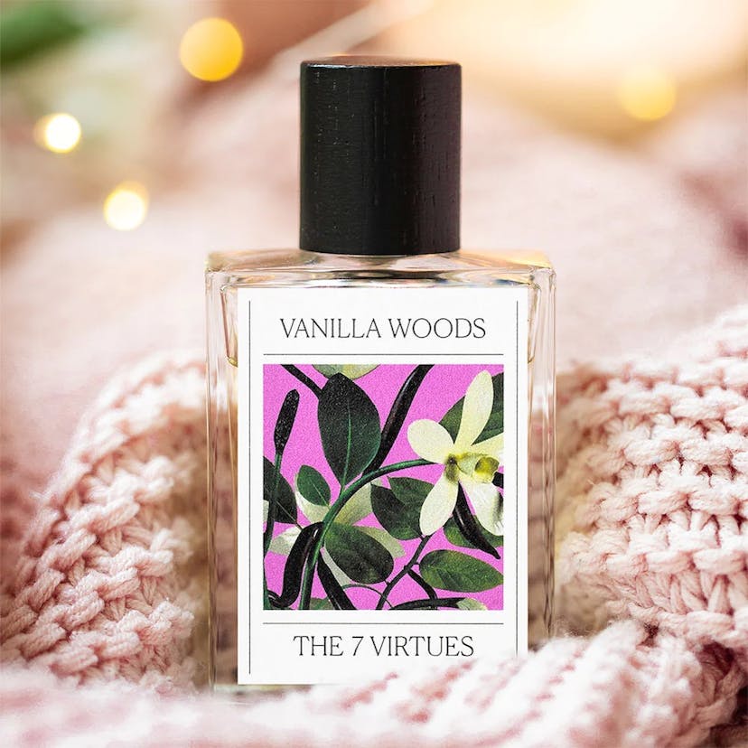 A clear, rectangular perfume bottle with a dark cap and a pink, floral label that reads “Vanilla Woods - The 7 Virtues” it atop a light pink knit blanket or scarf with diffuse twinkle lights in the background.