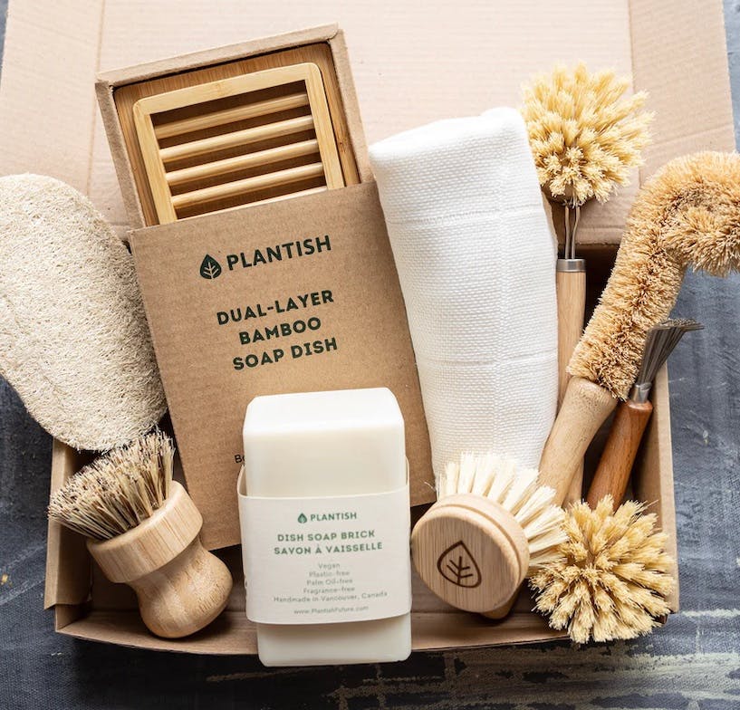 A cardboard box containing a dozen cleaning items for the kitchen, including several different scrub brushes, a soap brick, a soap dish, and a dish cloth.