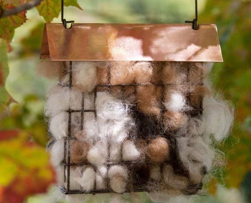 A small square cage bird feeder with a copper roof is filled with tufts of cream and brown-colored fur, shown hanging in front of green and red leaves.
