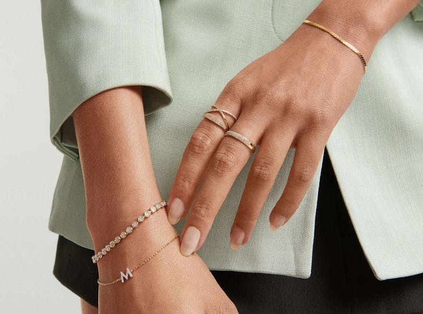 A woman’s hands are shown clasped in front of her seafoam green blazer and black skirt. On one hand, she wears a gold bracelet with a diamond letter M and a diamond tennis bracelet. On the other hand, she wears a thick, flat gold bracelet and several gold and diamond rings, all from jewelry company Mejuri.