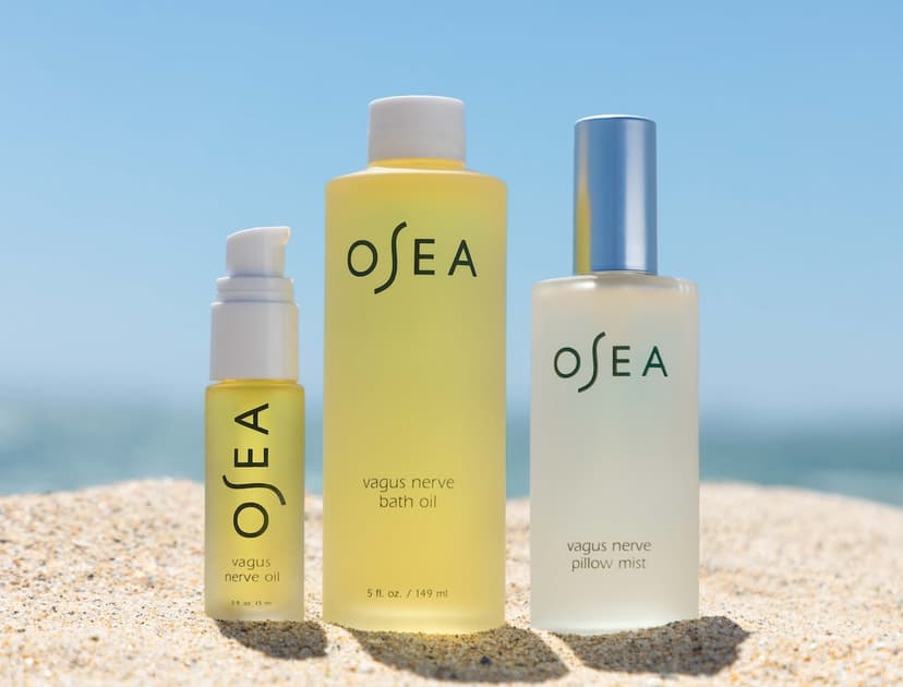 Three glass bottles sitting on sand on the beach, two containing yellow and one containing clear liquid. They are branded with the name OSEA. One reads "Vagus Nerve Oil," one reads "Vagus Nerve Bath Oil," one reads "Vagus Nerve Pillow Mist."
