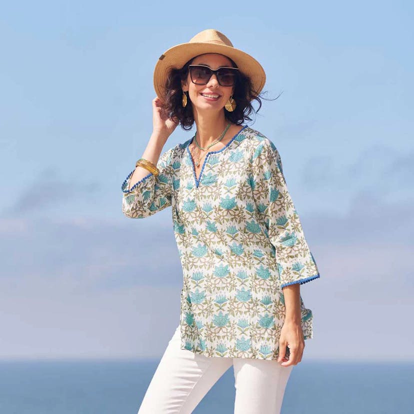 A smiling woman in sunglasses and a hat is wearing white pants and a beige and turquoise print top.