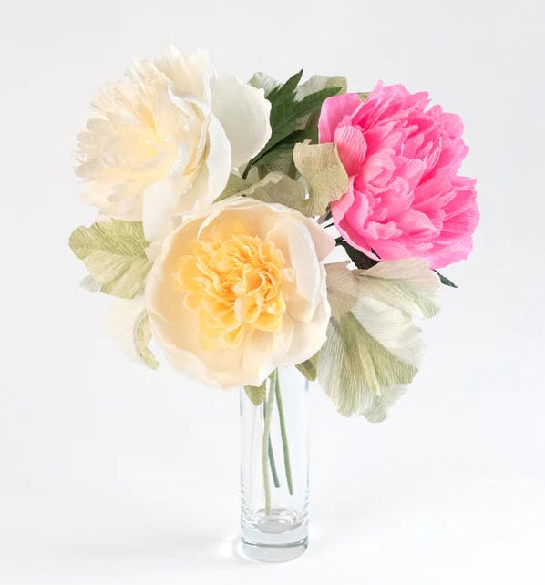 A bouquet of three peonies, one cream, one pale yellow, and one bright pink, all made from paper, in a clear glass vase.