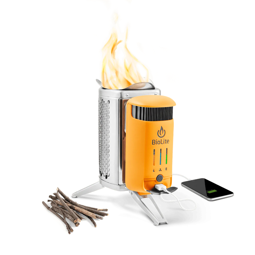 A small orange camp stove with the brand name BioLite on it is shown with flames coming out. Sticks rest on the side of the stove and a cell phone is plugged into the stove via USB cord.