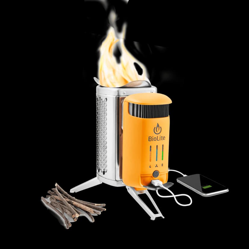 A small orange camp stove with the brand name BioLite on it is shown with flames coming out. Sticks rest on the side of the stove and a cell phone is plugged into the stove via USB cord.