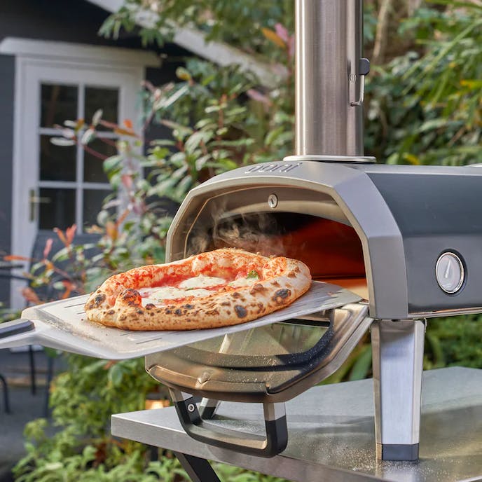A bubbling hot cheese pizza is being removed from a small silver and black Ooni pizza oven that is in shown in the backyard of a home.