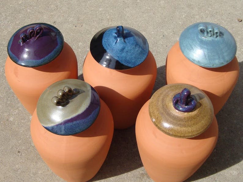 Five unglazed terra cot olla pots for passive watering. The pots are longer than they are tall, with attractive glazed ceramic tops in various hues of green, blue, and purple.