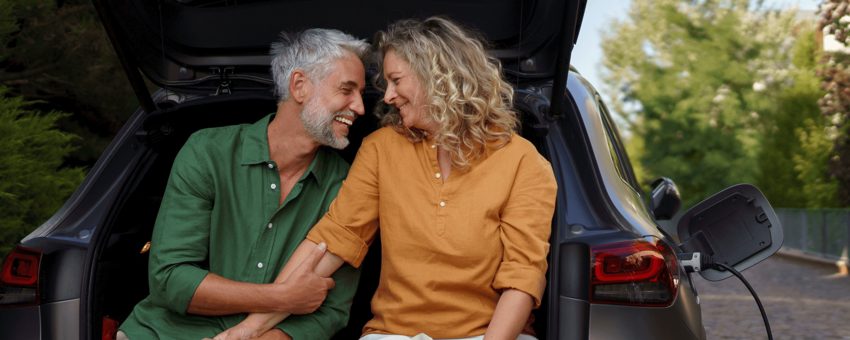 Smiling man and women sitting on the rear bumper of a charging electric car.