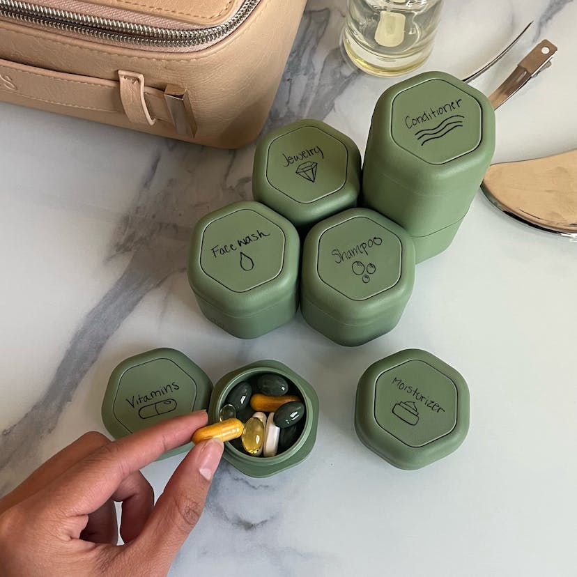 A white marble counter has seven opaque green hexagonal capsules on it labeled with the names of different products: Face Wash, Jewelry, Shampoo, Conditioner, and Moisturizer. Another capsule is ope and reveals vitamins and supplements inside, and a person’s hand is shown removing a vitamin pill.