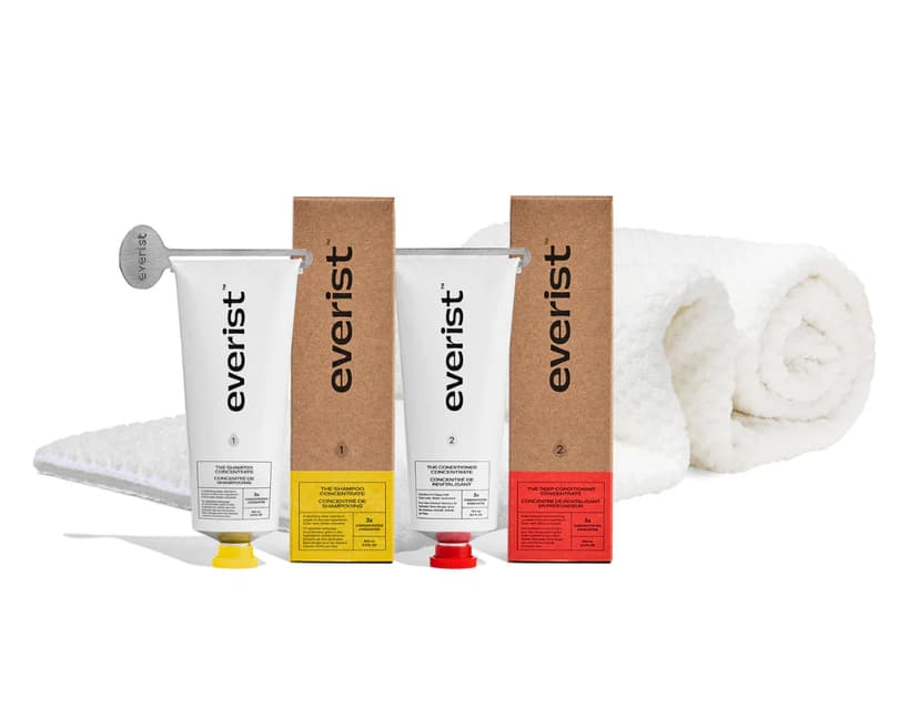 Two white tubes shown next to two cardboard boxes with a white towel in the background. The white tubes ach have a metal key at the top to help get all the product out of the tube. The products have the brand name Everist. The tube with the yellow top says “The Shampoo Concentrate” and the tube with the red top reads “The Conditioner Concentrate.”