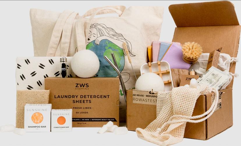 An assortment of items for sustainable living, some still in a cardboard box, including laundry sheets, wool laundry balls, a beige tote bag with a woman and the Earth on it, solid shampoo and conditioner bars, and more. A box of laundry sheets is labeled with the brand name ZWS Essentials