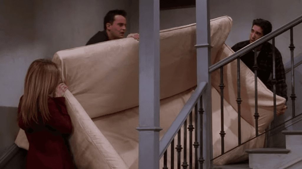 Three members of the cast of Friends moving a sofa up a staircase.