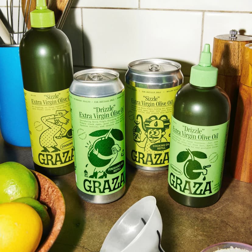 A cluttered kitchen counter with a blue bowl full of tools, a wooden bowl with lemons and limes, and two dark green plastic bottles and two silver aluminum cans, all with lime green labels for Graza Extra Virgin Olive Oil.