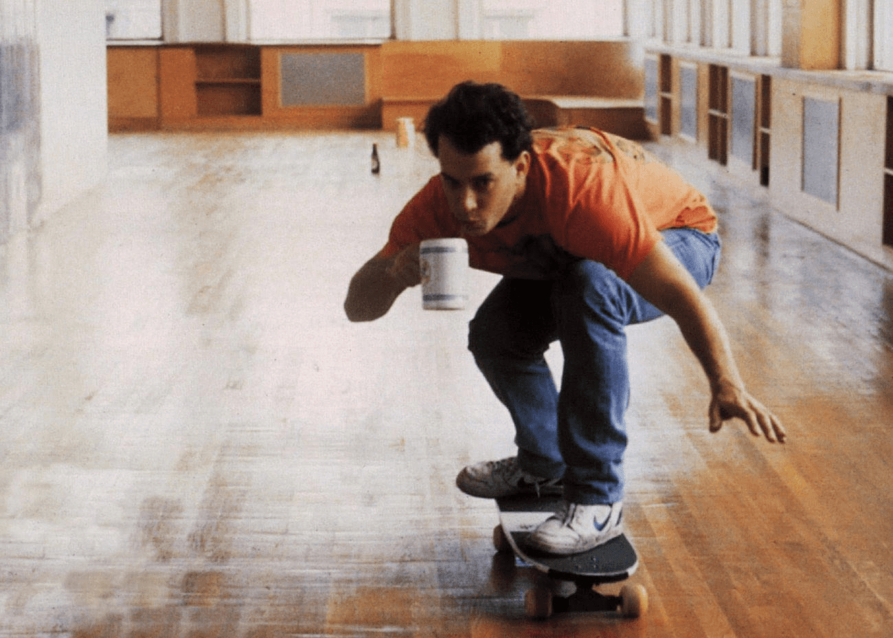 A man in his 20s or 30s rides a skateboard in a large open room and sips coffee from a mug.