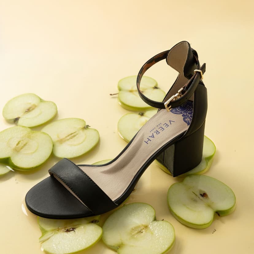 A strappy black block-heeled sandal with the brand name Veerah New York printed on the insole sits on top of sliced green apples.