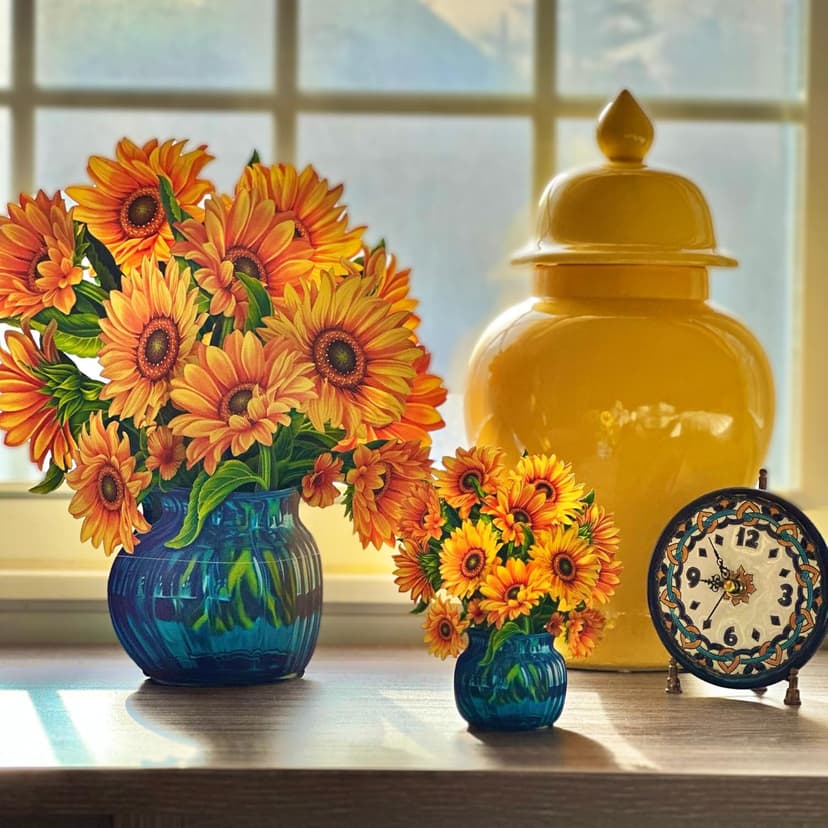 Two pop-up paper arrangements of sunflowers in blue vases, one small and one large, on a table in front of a window.