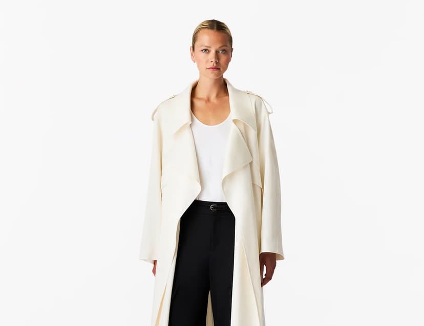A blond woman wears an elegant trench coat in an off-white color over a white scoop neck top and black pants, made by Another Tomorrow Apparel.