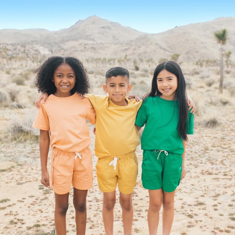 Three elementary-school-aged children stand in mountainous desert terrain wearing colorful matching shorts and tee-shirt sets, one in orange, one in yellow, and one in green.