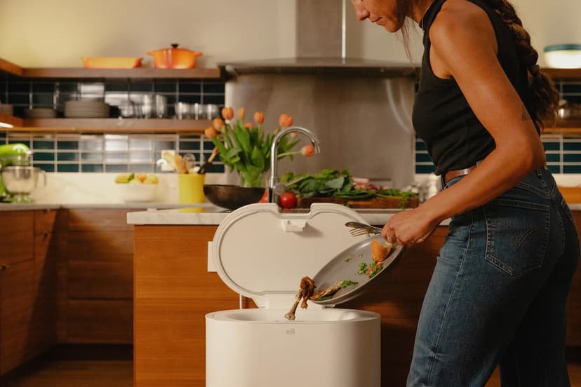 A woman in a black tank top and blue jeans stands in a modern kitchen, scraping food scraps off a plate into a white bin with the brand name Mill. The kitchen features wooden cabinets, a marble countertop, and a variety of fresh vegetables and fruit in the background. A bouquet of orange tulips sits on the counter, and a mixer is visible nearby.