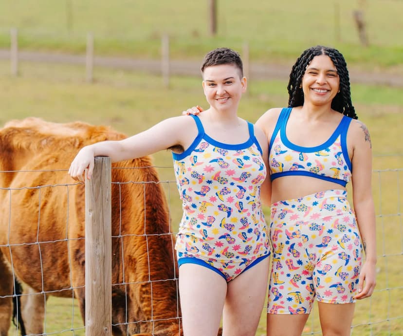 Two women wearing cowboy-print underthings standing in front of a brown cow