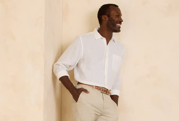 A relaxed-looking man smiles, wearing a pair of khaki trousers and a cream-colored linen button-up shirt from Tommy Bahama.
