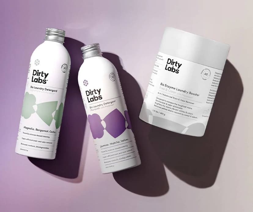 Two white bottles with green and purple markings and a white and grey cylindrical package, all labeled as Dirty Labs brand Bio Laundry cleaning products, on a lavender and beige-colored background.