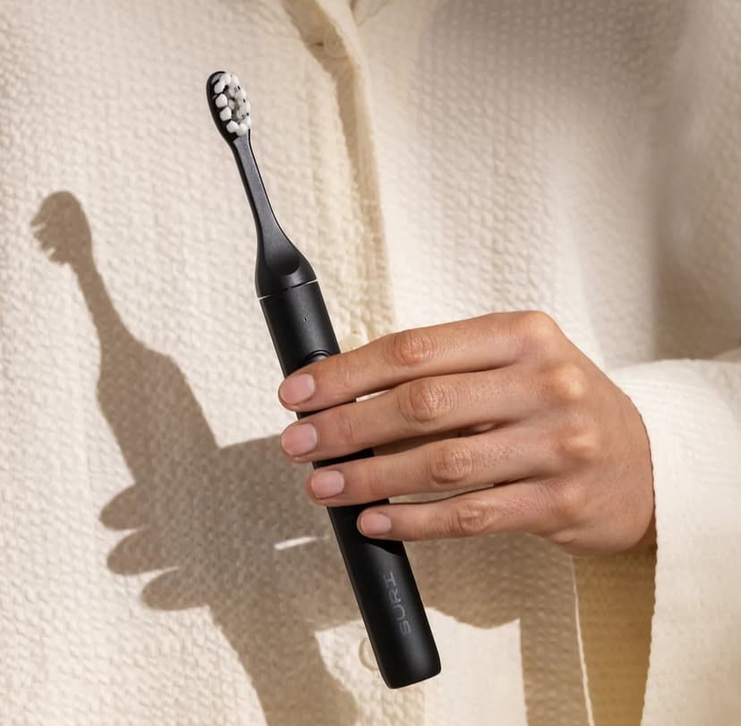 A close-up on the torso of a person wearing a cream-colored seersucker blouse, holding a slim black electric toothbrush stamped with the brand name SURI.