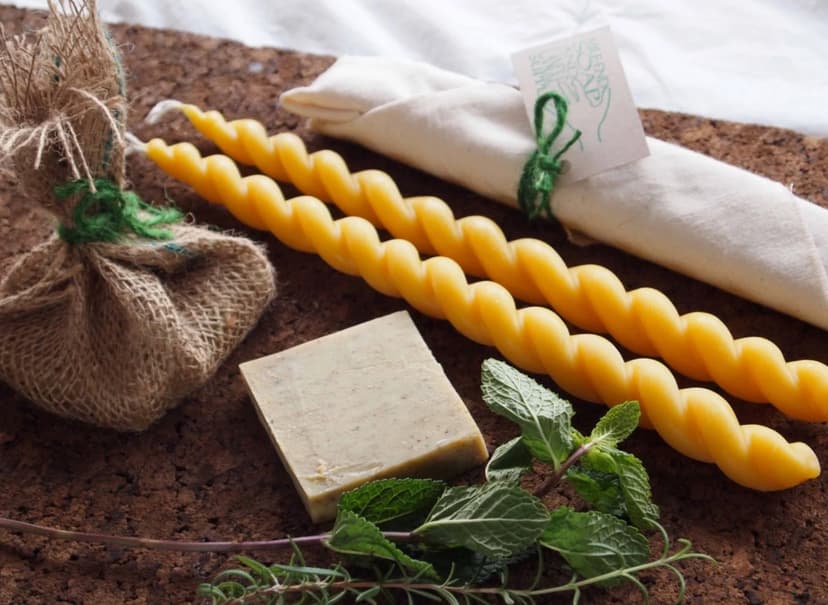Two long and twisty beeswax candles, a small square beige soap, a small burlap package tied with green string, and a small wrapped packaged with a card reading Mile End Soap and Candle Supply are displayed alongside fresh rosemary and mint on a marble countertop.