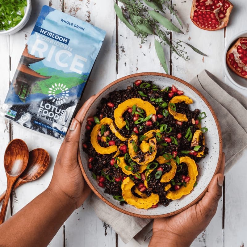A person’s hands cradle a bowl of cooked dark-purple rice with sliced rings of squash, pomegranate seeds, and green onions. In the background, a back of Lotus Foods Heirloom Forbidden Rice.