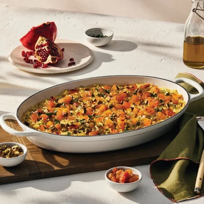 A white oval baking dish with a squash casserole inside, with garnishes on the side, including a pomegranate.