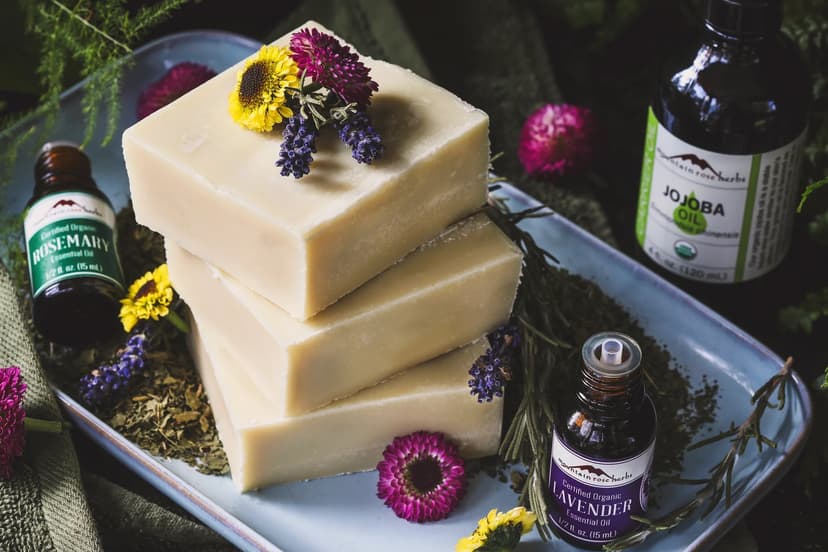 Three bars of soap on a plate, surrounded by bottles of essential oils from the brand Mountain Rose Herbs and colorful fresh flowers.