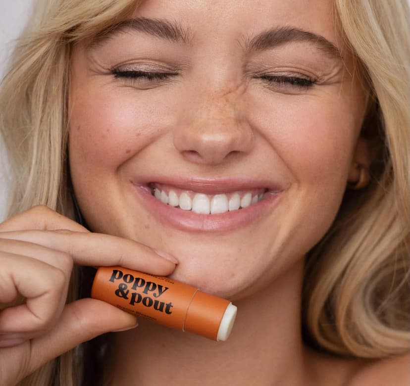a blonde woman smiles and holds up an orange lipstick tube with the brand name Poppy & Pout printed on it