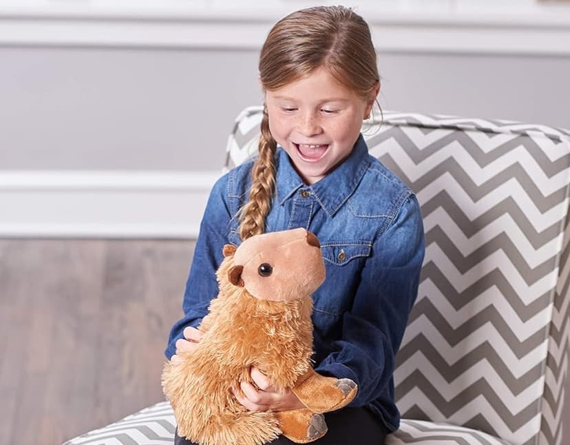 A strawberry girl in a denim shirt sitting on a grey and white striped chair smiles broadly as she looks down at the brown Capybara Wild Republic stuffed animal toy on her lap.