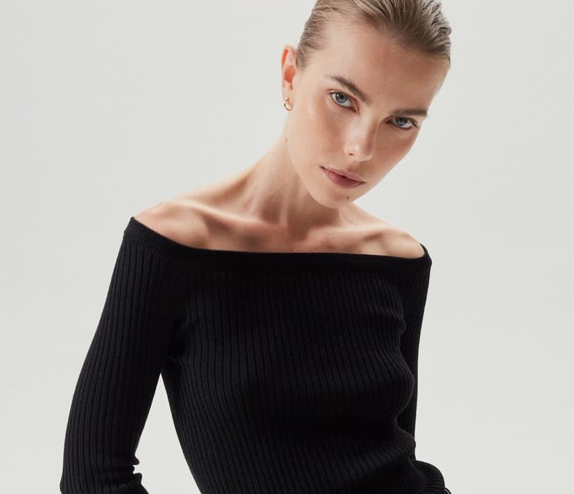A woman with slicked back blonde hair and blue eyes wears a ribbed, off-the-shoulder black top.