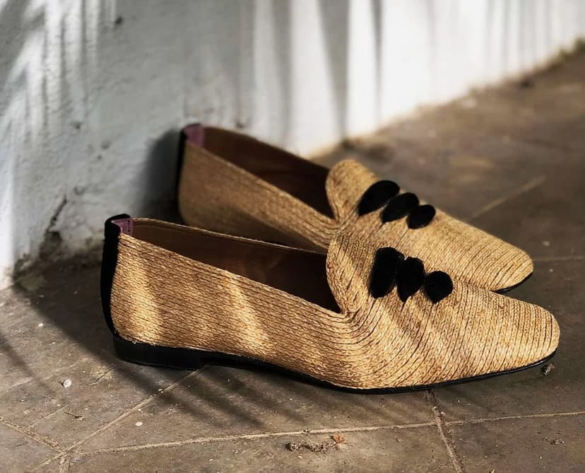 A pair of women’s raffia flat shoes with black laces in an outdoor corridor with whitewashed walls and palm frond shadows.
